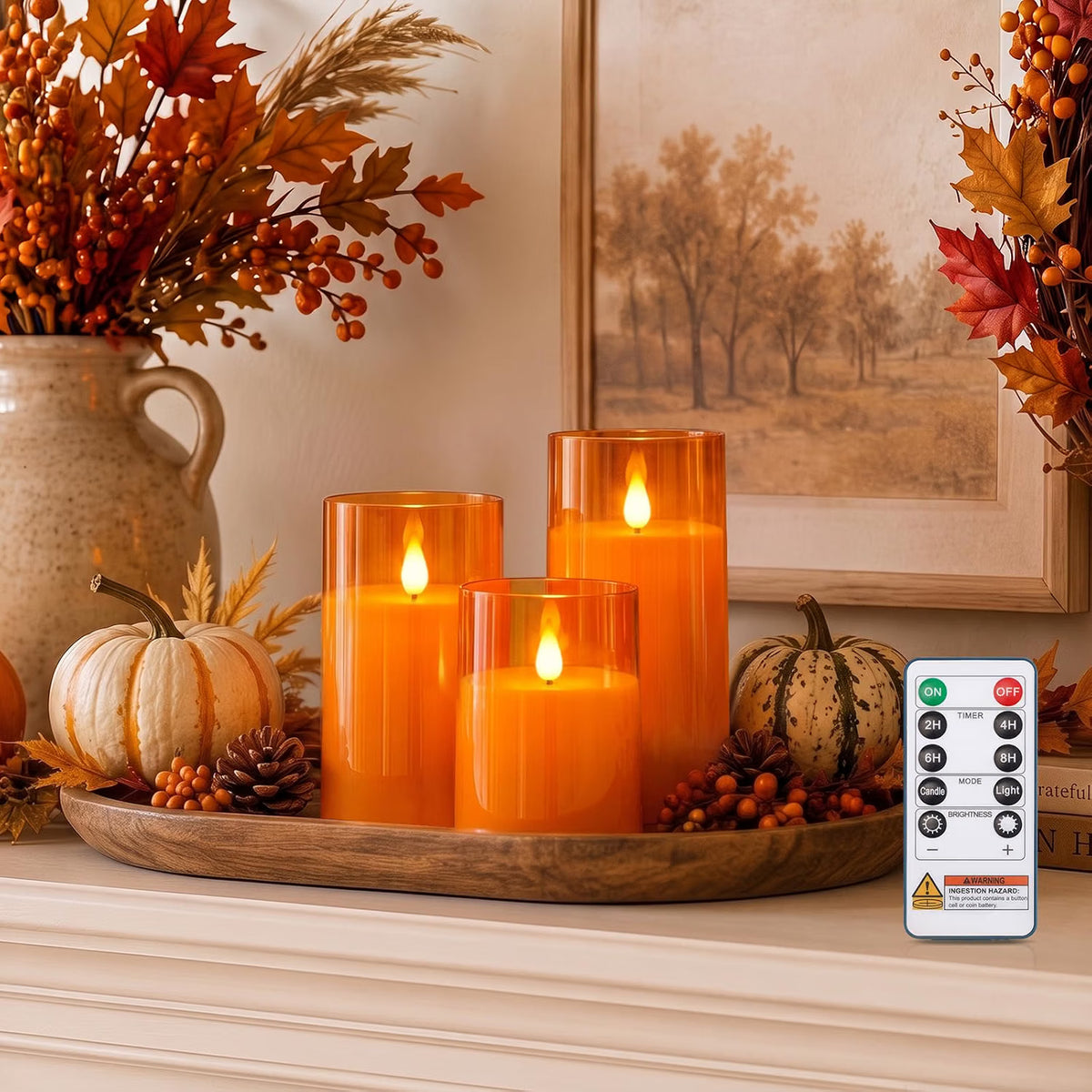 Three orange pillar candles with flame-shaped lights on a wooden tray, styled with pumpkins, berries, and fall decor, with a white remote control shown in the lower right corner.