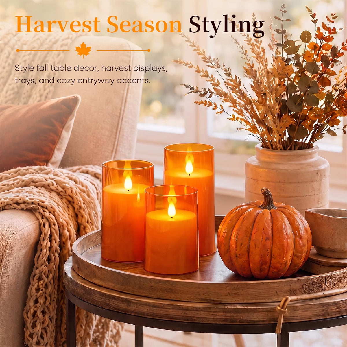 Three flameless orange glass pillar candles displayed on a round wooden tray in a cozy fall living room setting, alongside a small orange pumpkin, a knitted throw blanket, and a vase of dried botanicals. Text at the top reads 'Harvest Season Styling'.